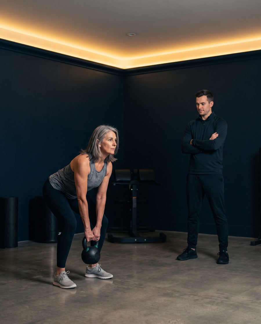 A gray-haired woman lifting a kettlebell in a training studio while a coach watches her form